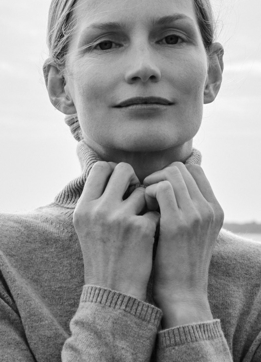 A close-up black and white portrait of a woman wearing a ribbed turtleneck sweater, with her hands adjusting the collar.