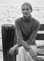 Model Katrin Thormann seated by the water wearing light grey cashmere rollneck jumper and ivory skirt